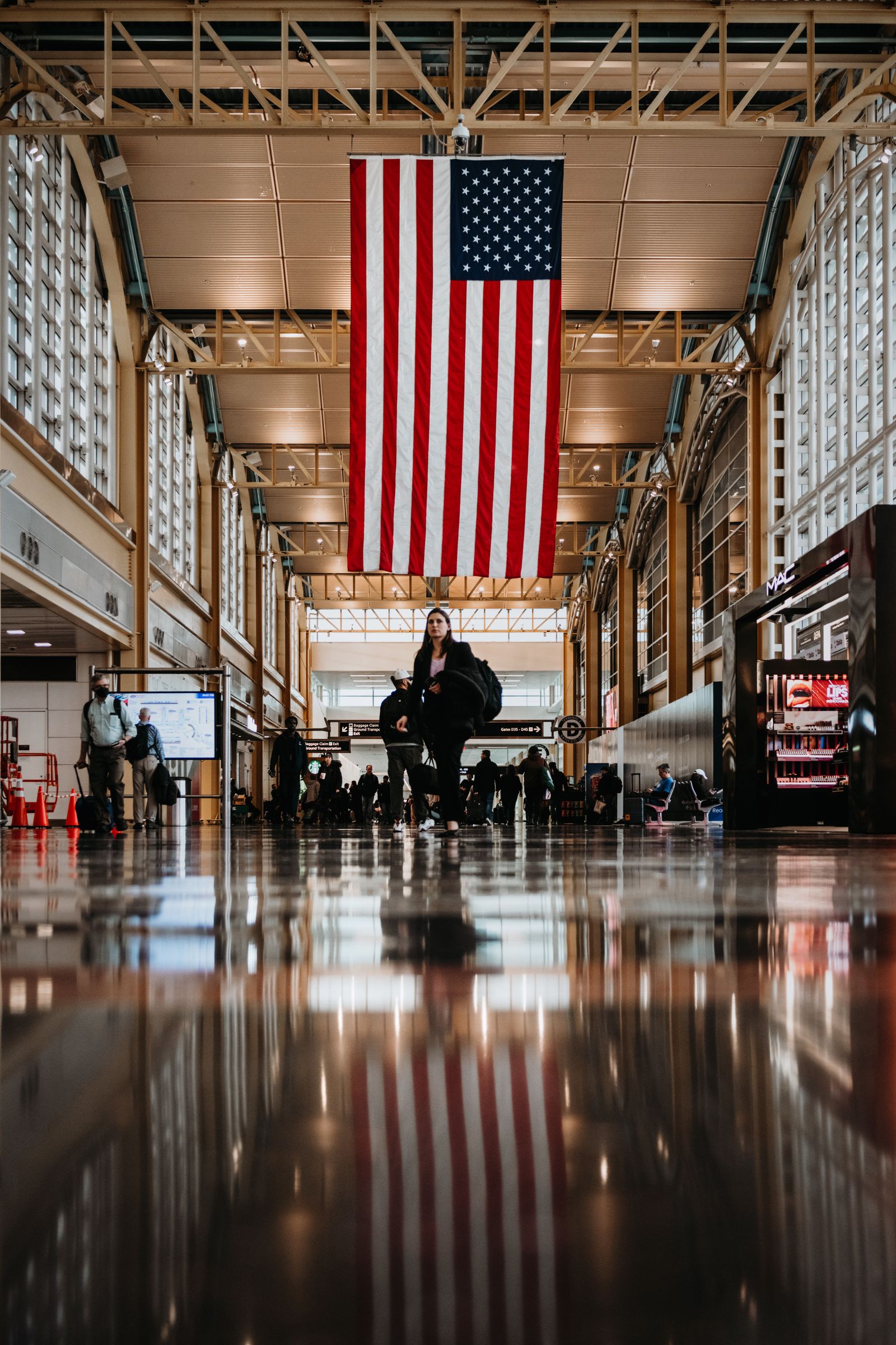 Viajantes no interior do Aeroporto Nacional Reagan, com a bandeira dos Estados Unidos ao fundo