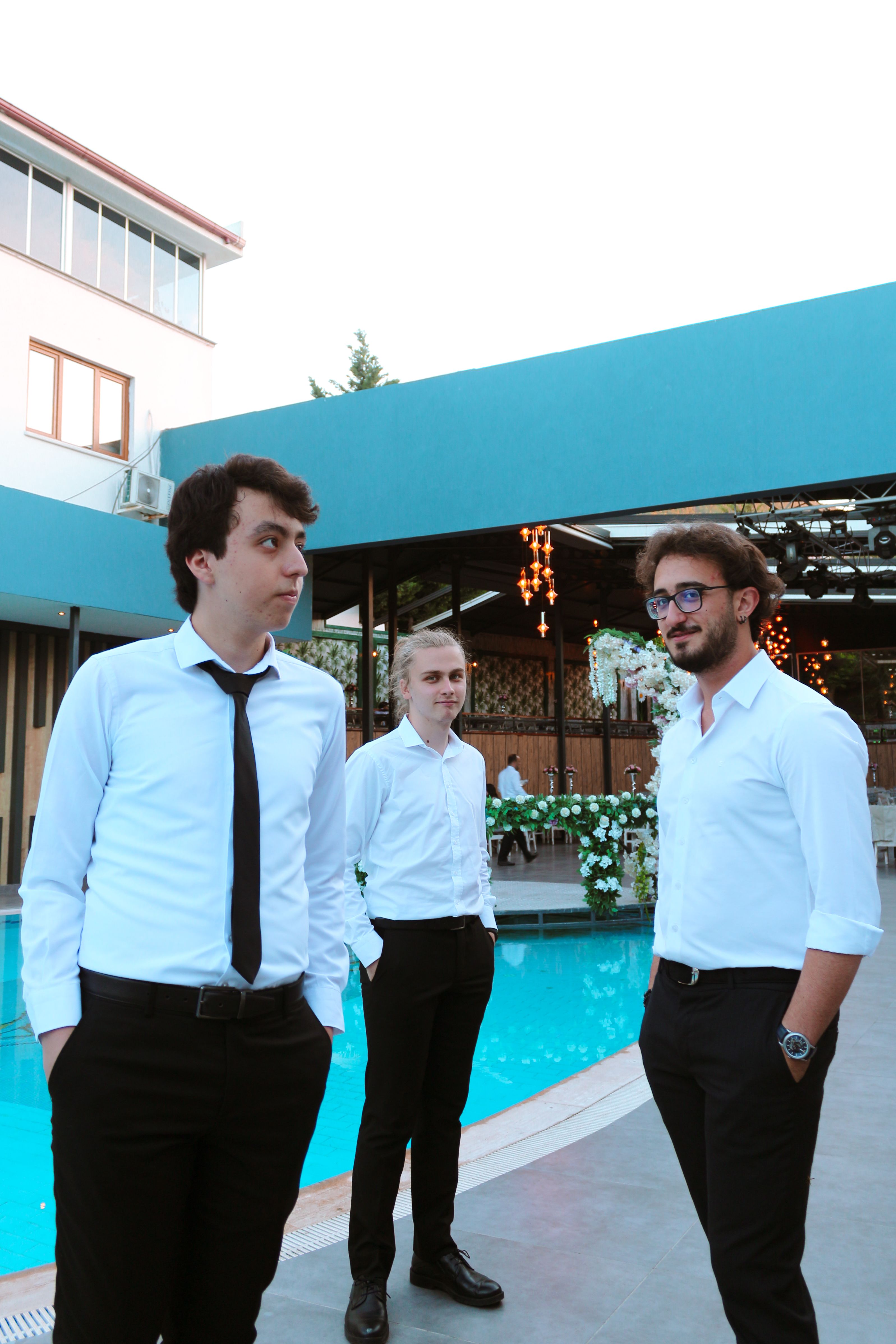 Three men in formal attire standing together at an outdoor venue near a pool.