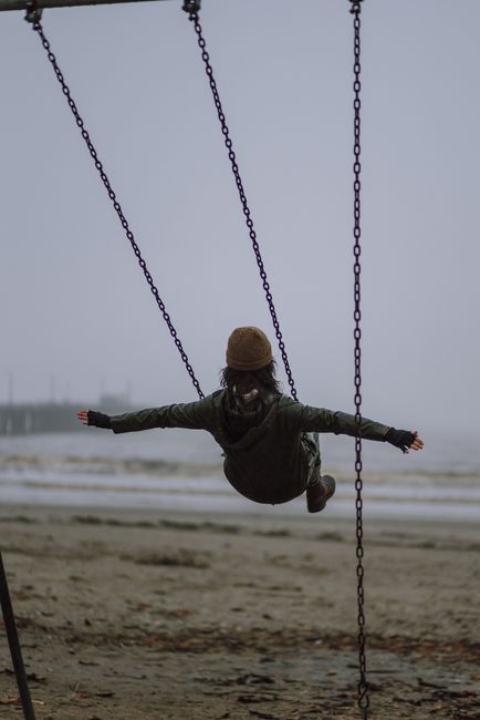 A woman in warm clothing swings joyfully on a foggy beach, capturing a serene and playful moment.