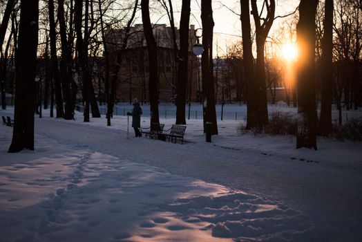 A tranquil park covered in snow as the sun sets, highlighting benches and footprints.