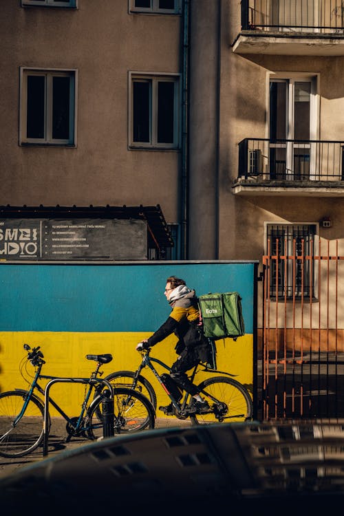A Food Delivery Man on a Bicycle Riding near a Residential Building and a Wall with a Ukrainian Flag 