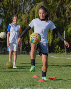 Two girls in sports attire practicing soccer drills on a sunny day outdoor field.