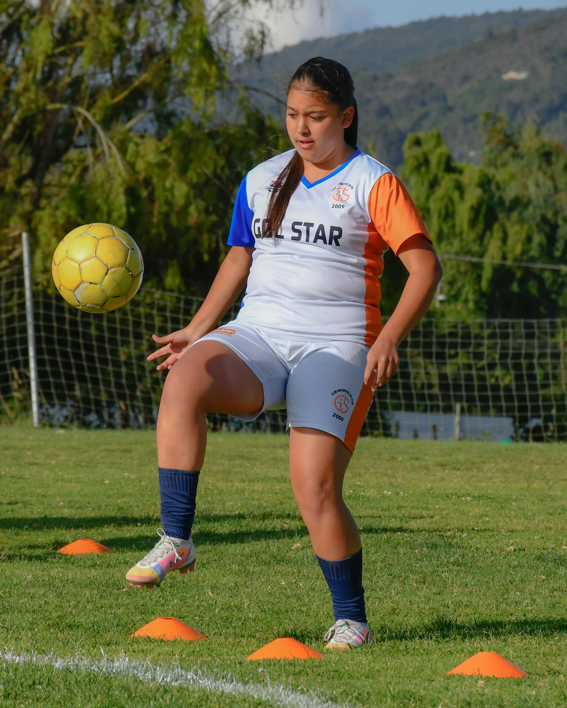 A girl in sportswear practices soccer ball control on a green field during the day.