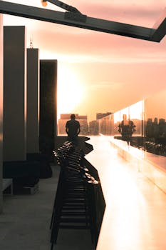 A man stands on a rooftop with a city skyline view during sunset, creating a serene urban scene.