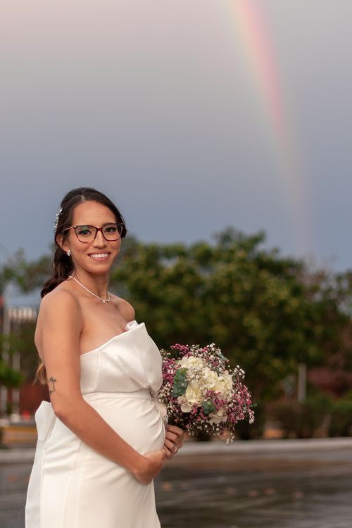 Free A smiling pregnant bride in a wedding dress holding a bouquet with a rainbow in the background. Stock Photo