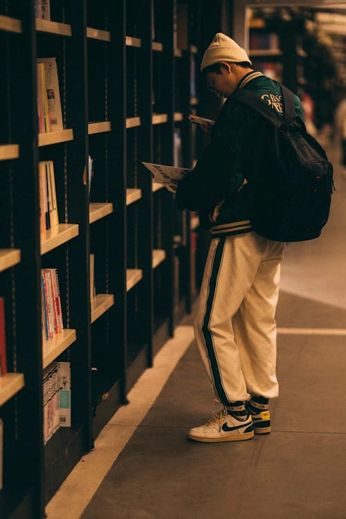 A College Student in a Library Standing next to a Shelf and Holding a Book 