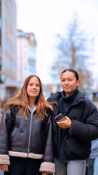 Two young women standing on a city street wearing winter coats, smiling at the camera.
