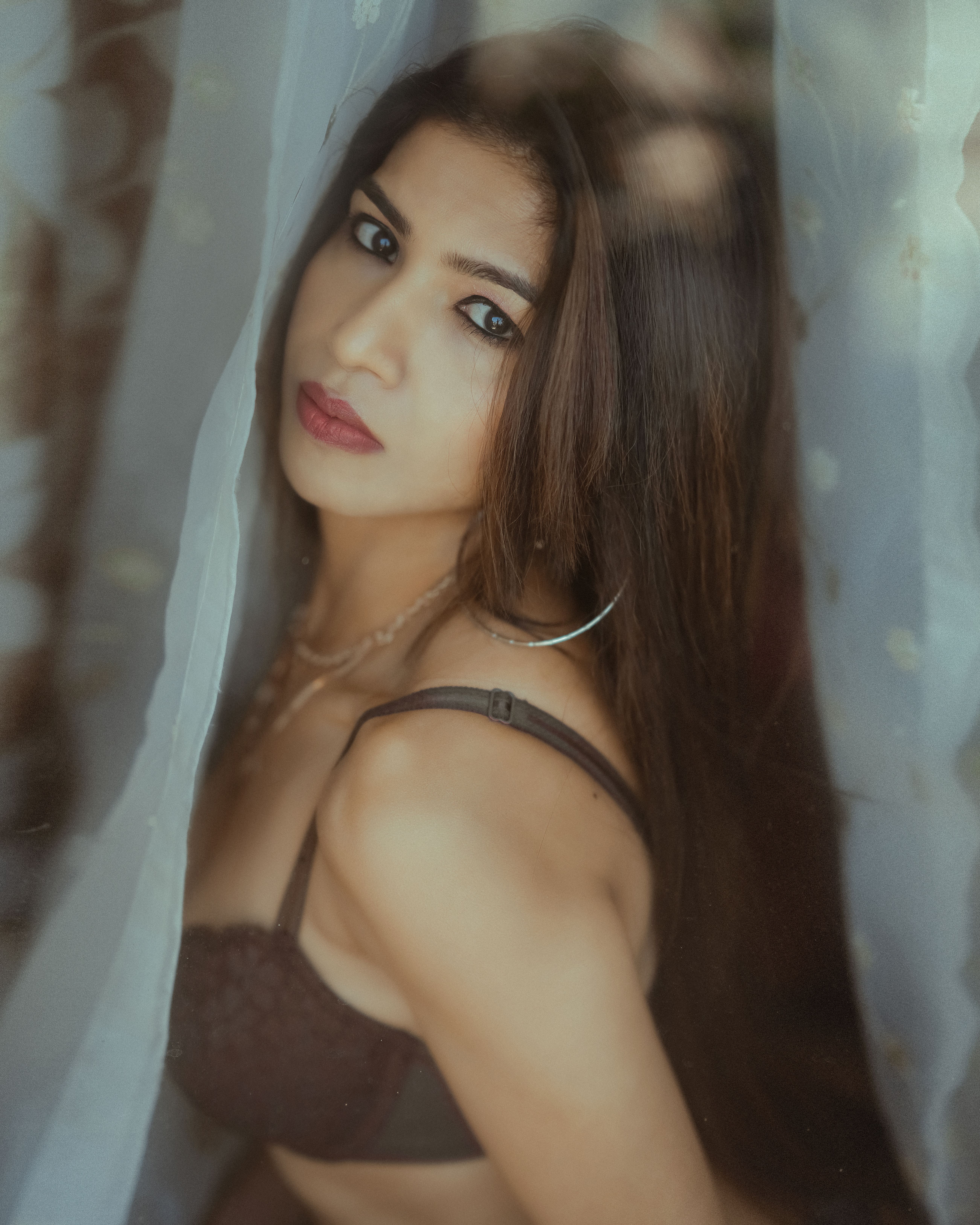 A captivating portrait of a brunette woman in lingerie, posing behind sheer curtains. The mood is soft and elegant.