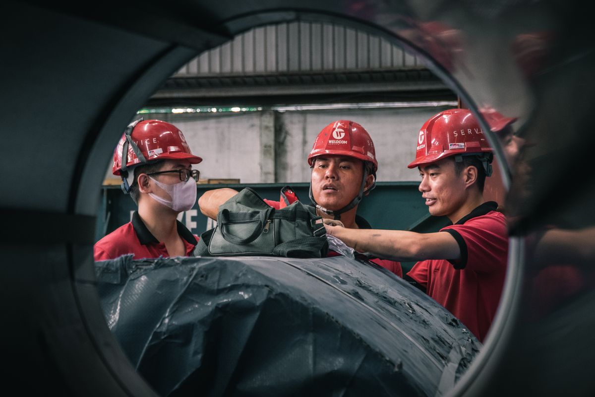 Three Southeast Asian workers in safety helmets discussing work on site, the pre-shift briefing where night lighting arrangements are confirmed