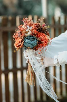 A close-up of a rustic bridal bouquet with colorful flowers and lace ribbon, held by a bride outdoors.
