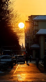 A city street bustling with cars and people under a striking sunrise, casting silhouettes and warm tones.