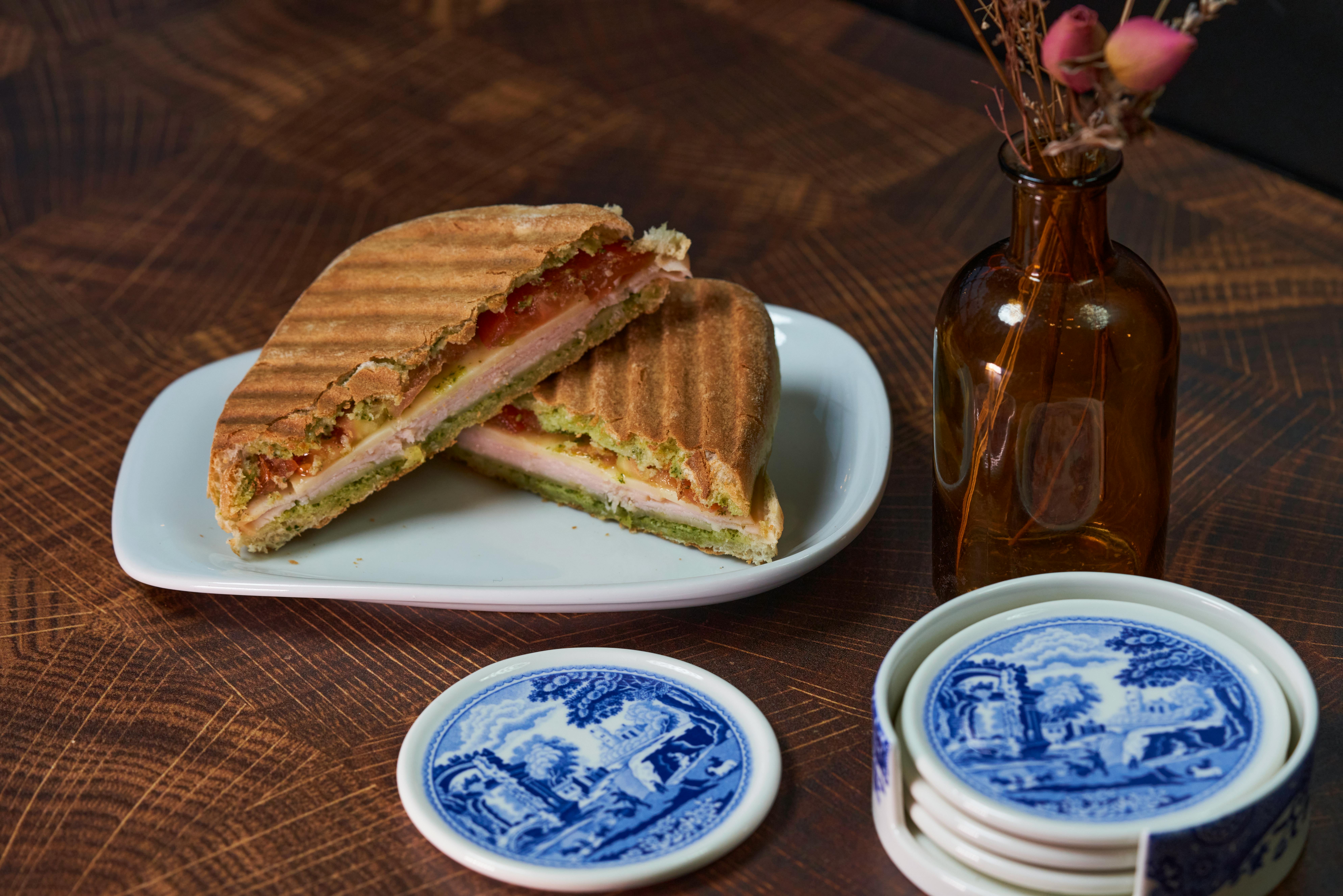 Delicious panini sandwich served on a white plate, with artistic blue coasters and a decorative vase on a wooden table.