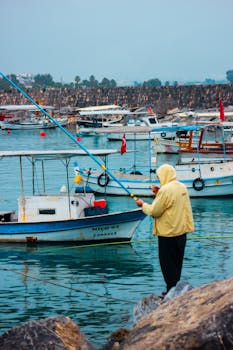 A man fishing by the coast in Dörtyol, Hatay, Türkiye. Boats anchored in the peaceful harbor.