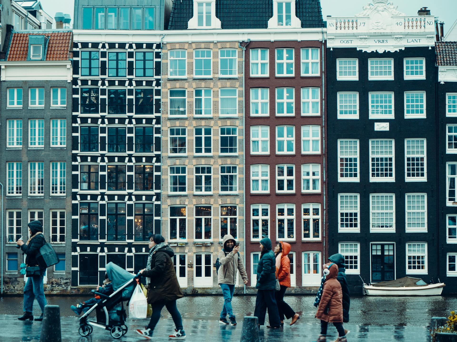 People walking in rain by iconic Amsterdam canal houses, showcasing urban life.