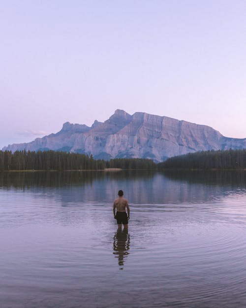 Back View of Man Standing in the Two Jack Lake with View of a Forest and Mountains 