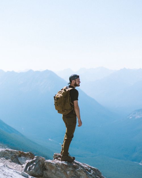 Free A man with a backpack stands atop a rocky mountain peak, embracing the expansive valley view. Stock Photo