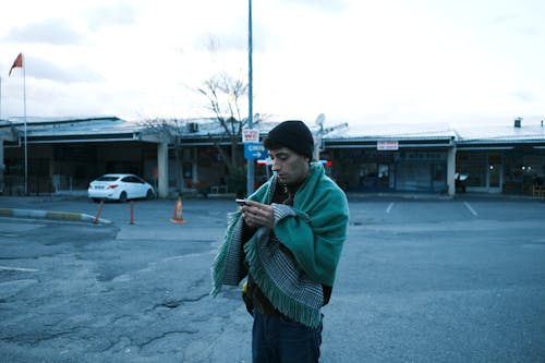 Man Wearing a Green Shawl Standing in a Parking Lot with a Smart Phone in Hands