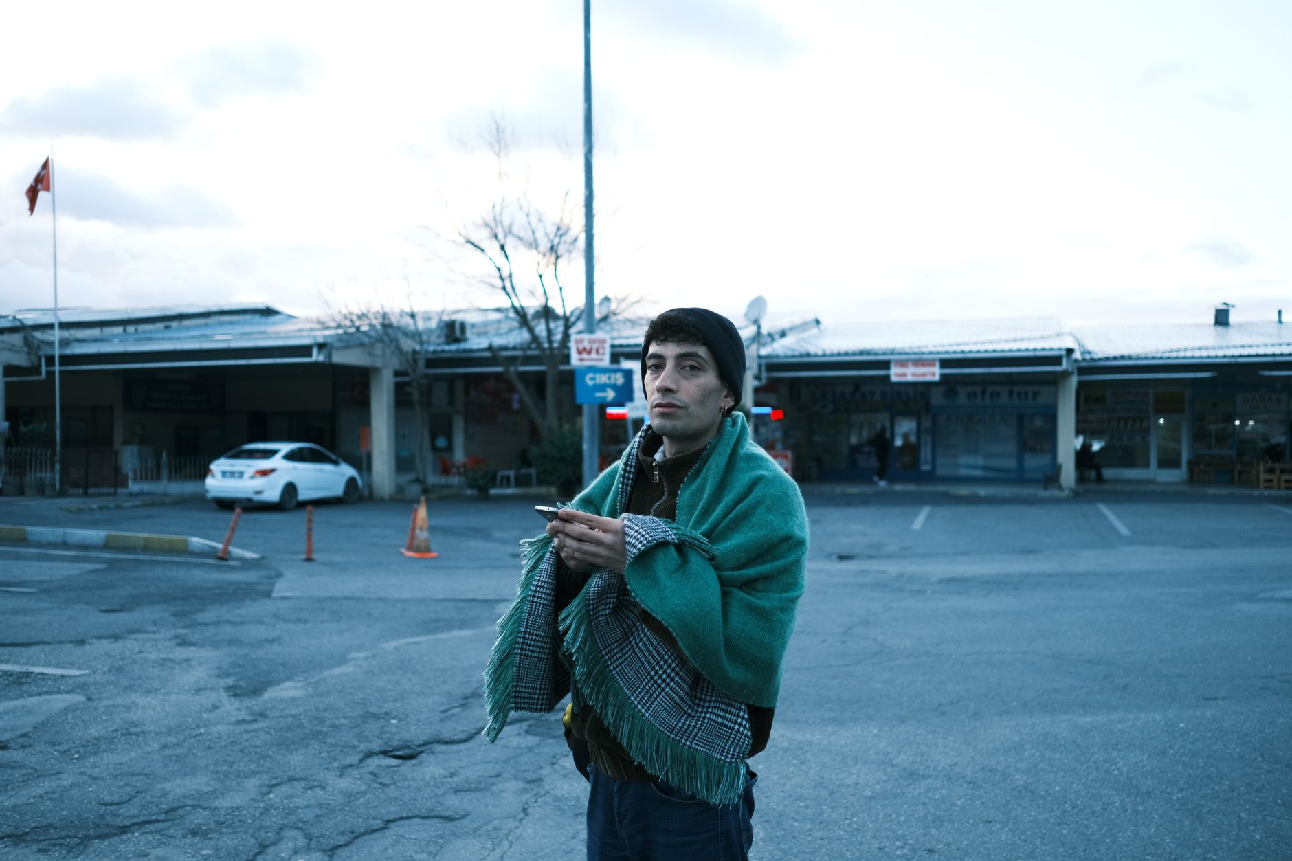極地旅行手機電池低溫保暖:衛星通訊救急指南 A young man stands wrapped in a blanket, holding a phone at a parking lot during dawn.