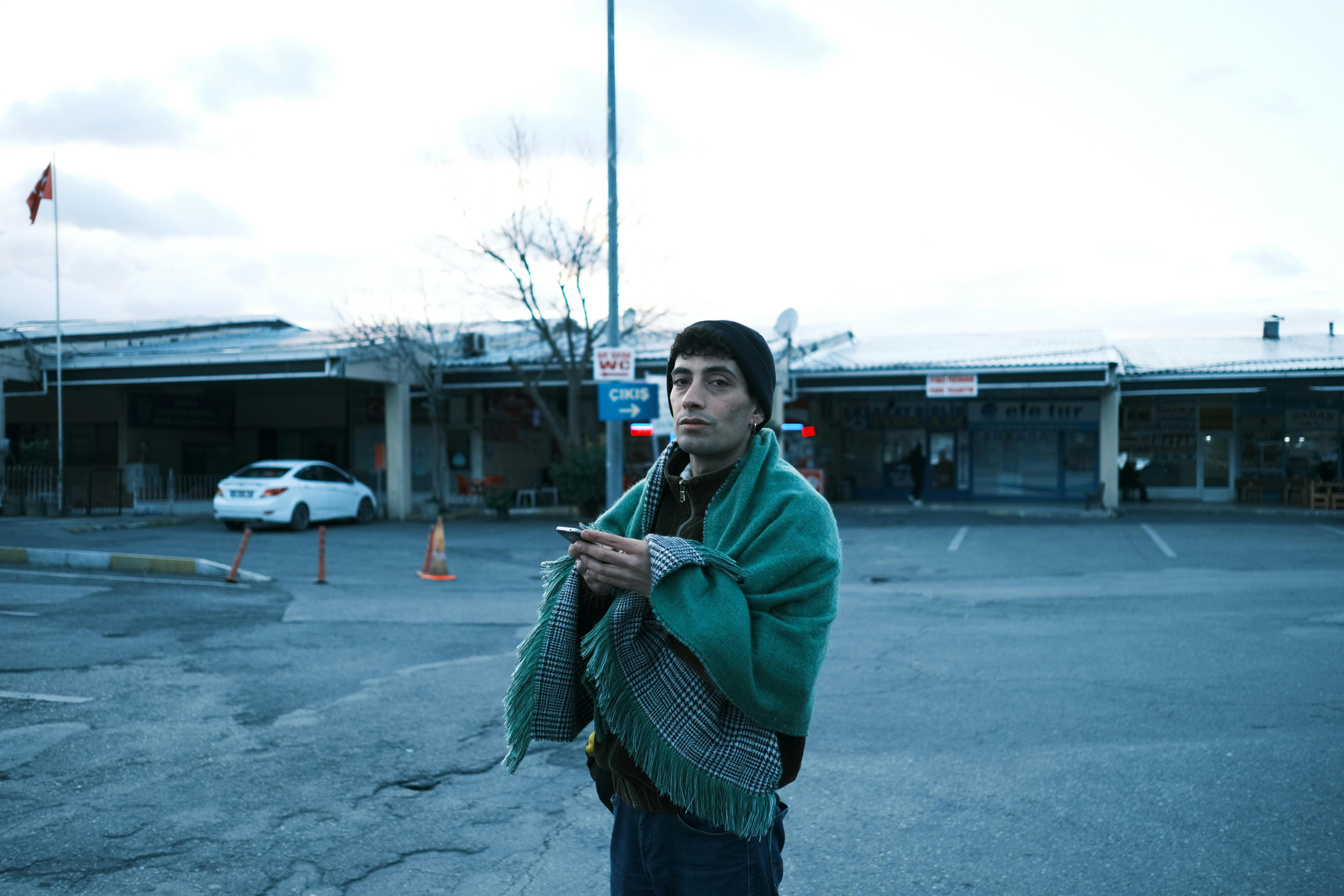 A young man stands wrapped in a blanket, holding a phone at a parking lot during dawn.