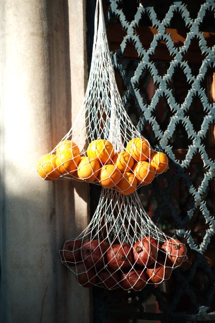 Net bags of oranges and pomegranates hanging on a textured iron wall.