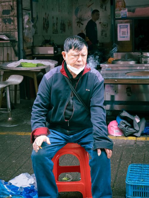 Free Asian man sitting outdoors with mask, surrounded by market items, smoking. Stock Photo