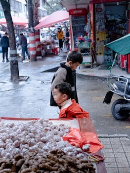 Two children observe a bustling street market stall with garlic and ginger on display.