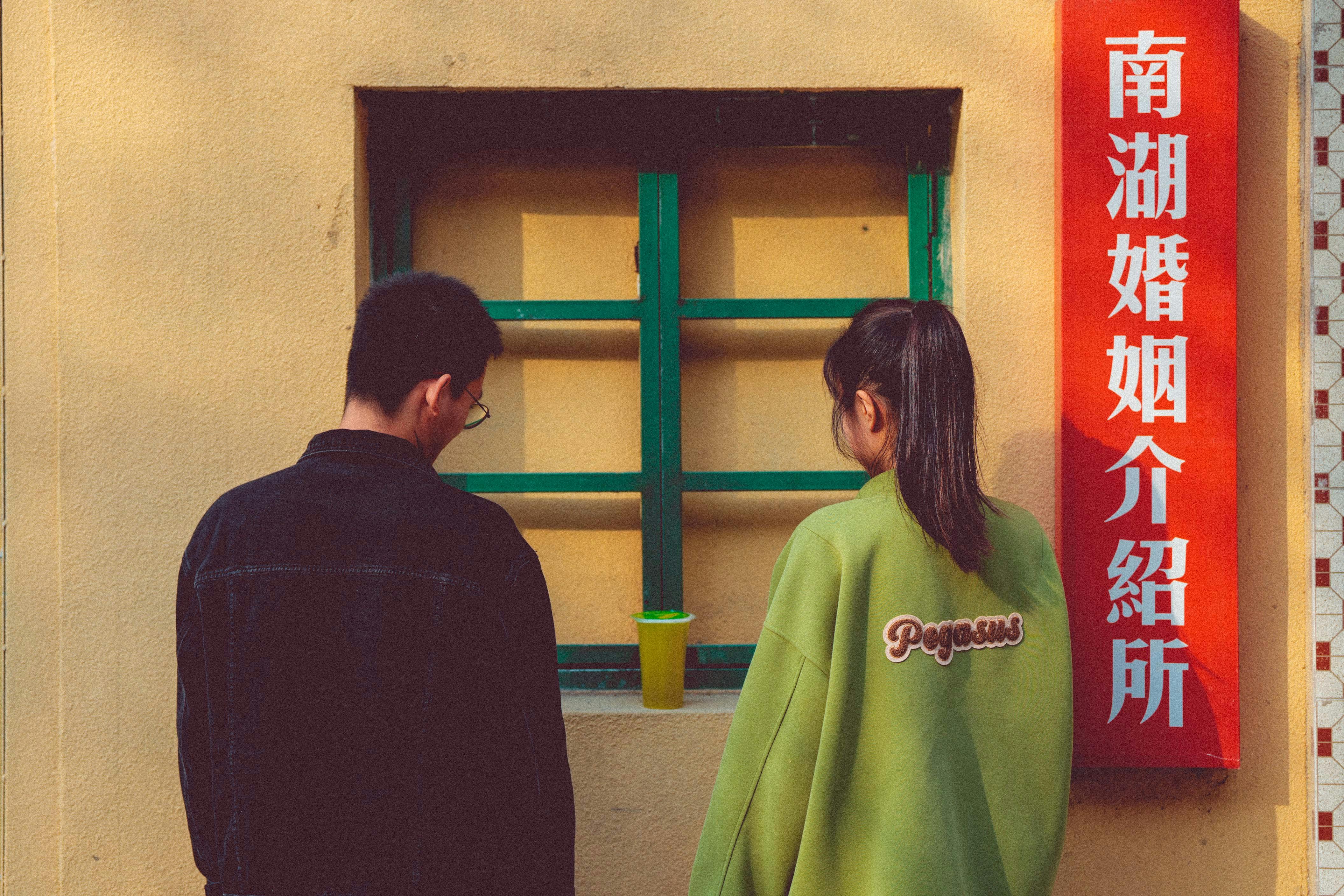 A man and woman stand side by side by a window with Asian text and vibrant wall.