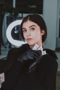 Elegant woman in black gloves holding cosmetic products, poised against a ring light backdrop.