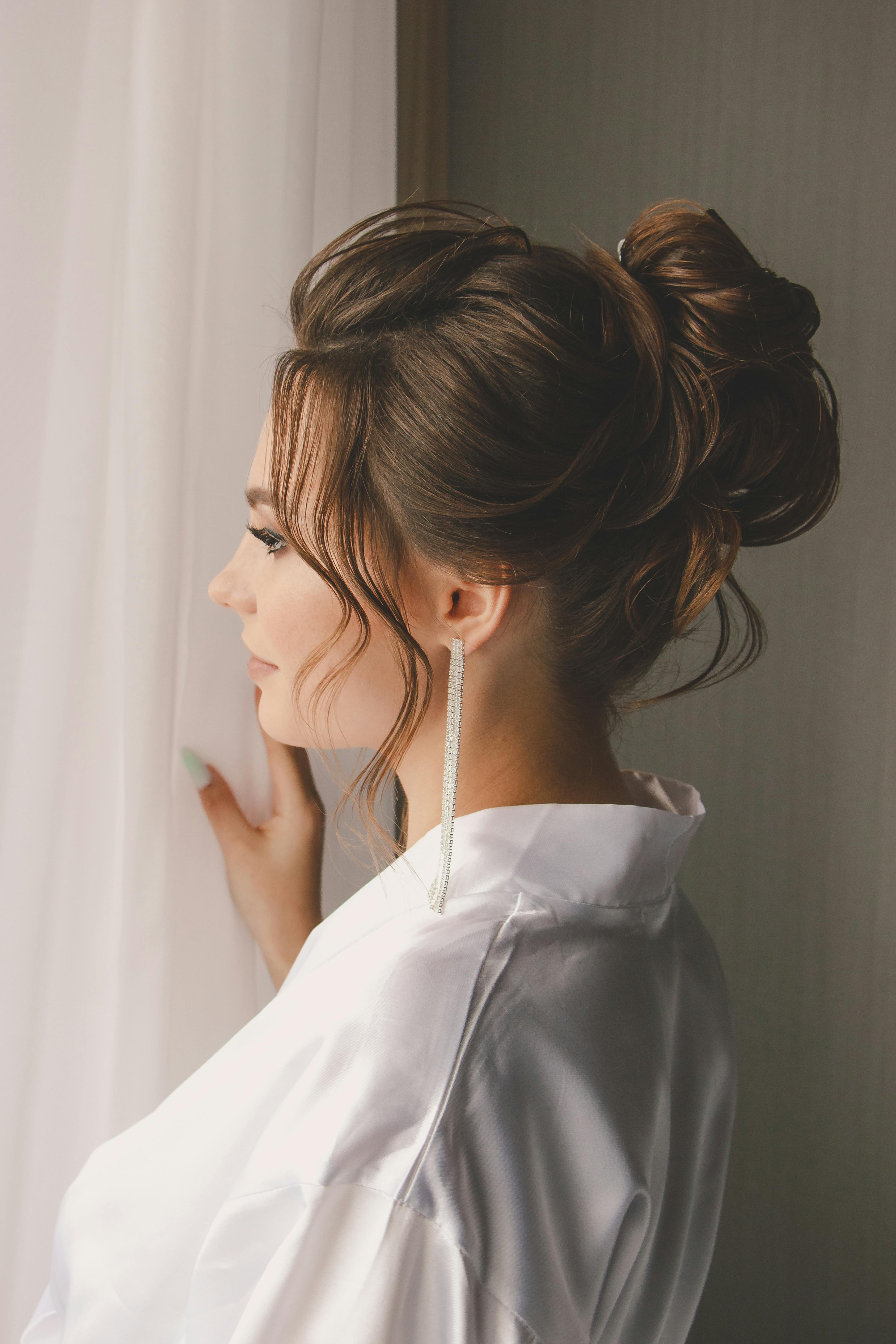 A young woman with an elegant hairstyle poses in a white shirt by the window.