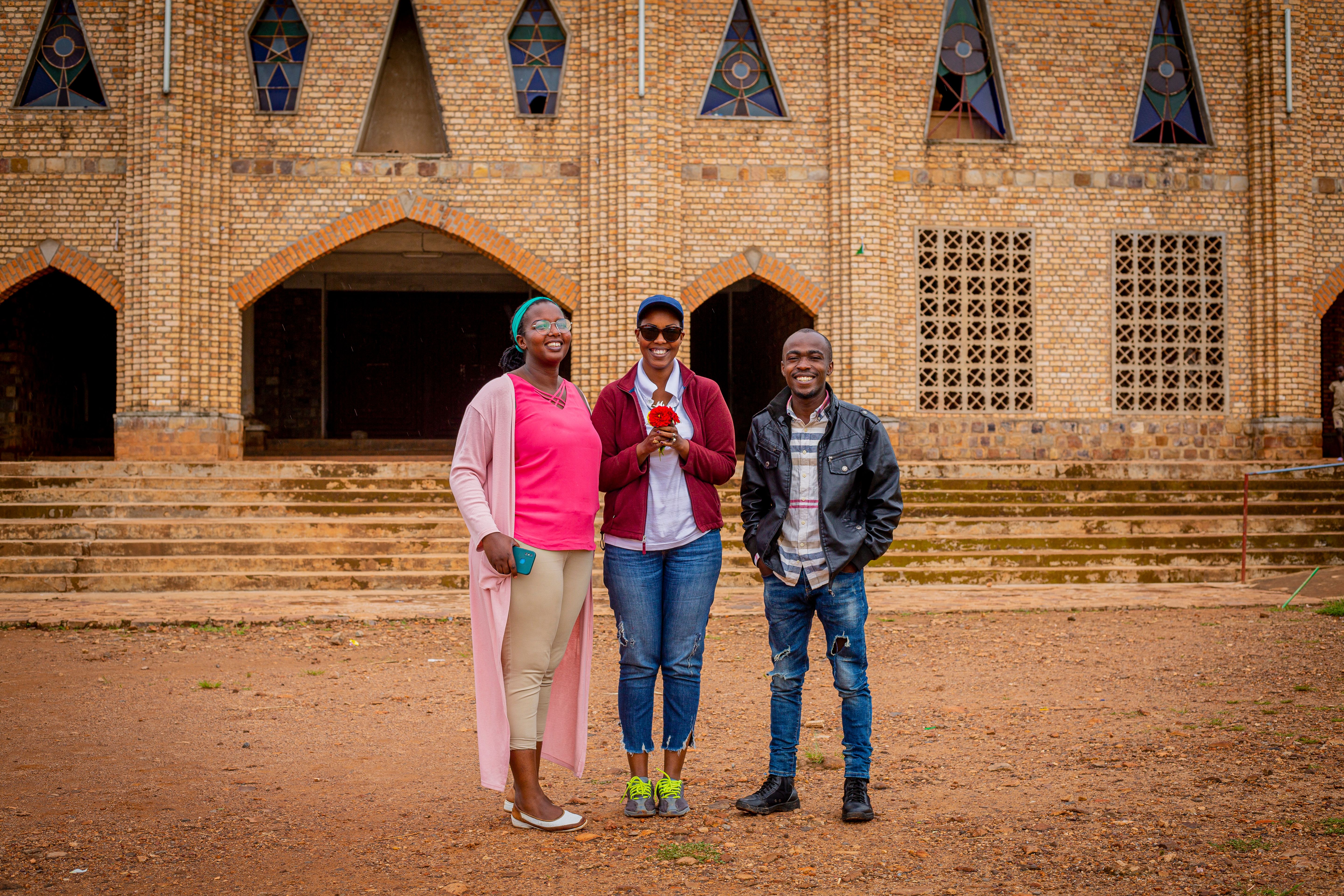 Three friends stand smiling in front of a brick church facade, enjoying a sunny day.