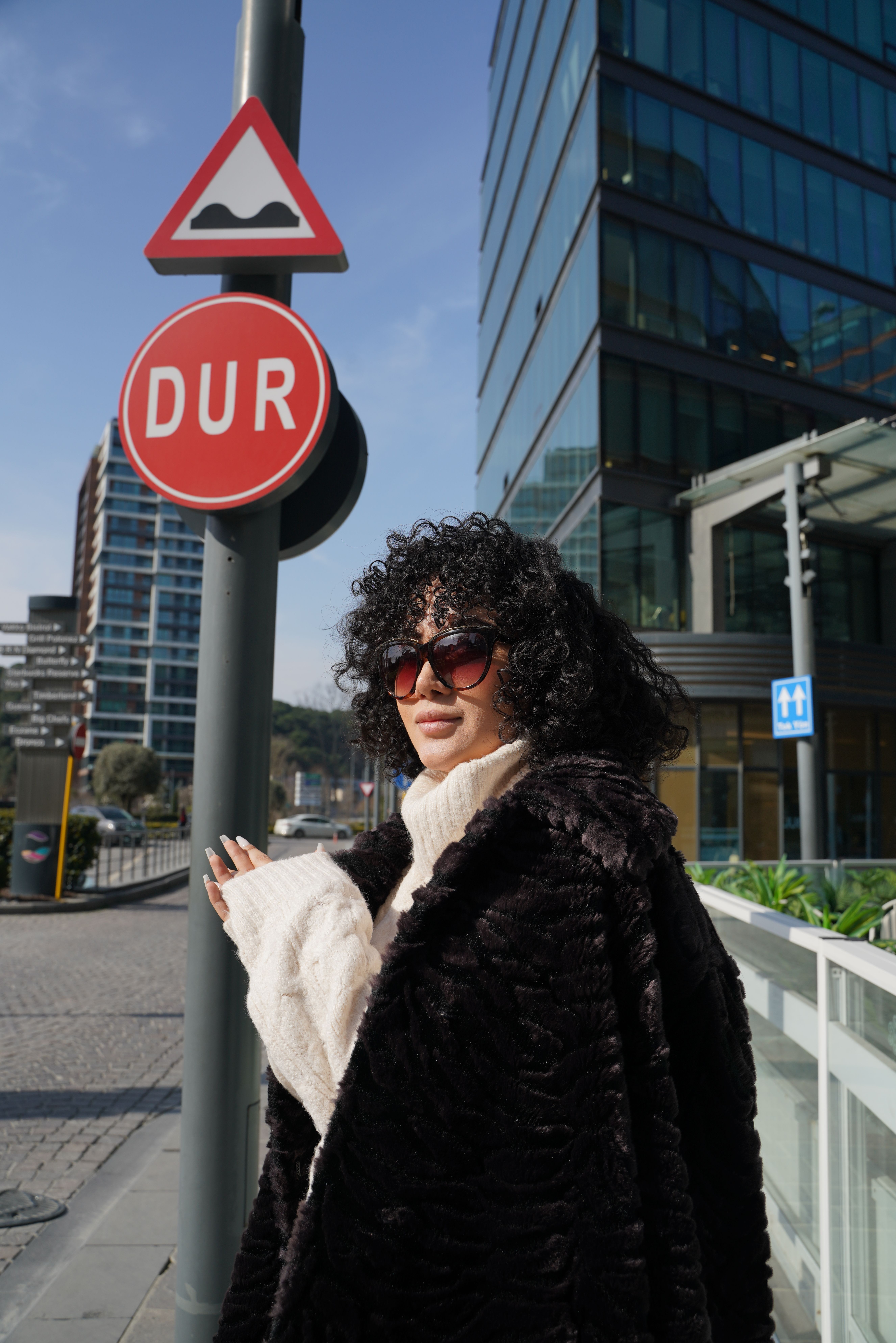 Stylish woman posing in urban İstanbul with 'Dur' sign and modern architecture.