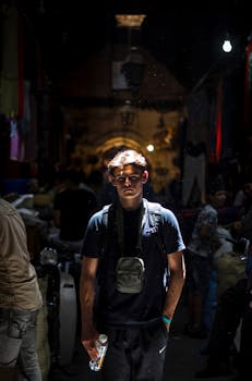 A young man stands confidently in a dimly lit Marrakech market, with vibrant fabrics surrounding him.