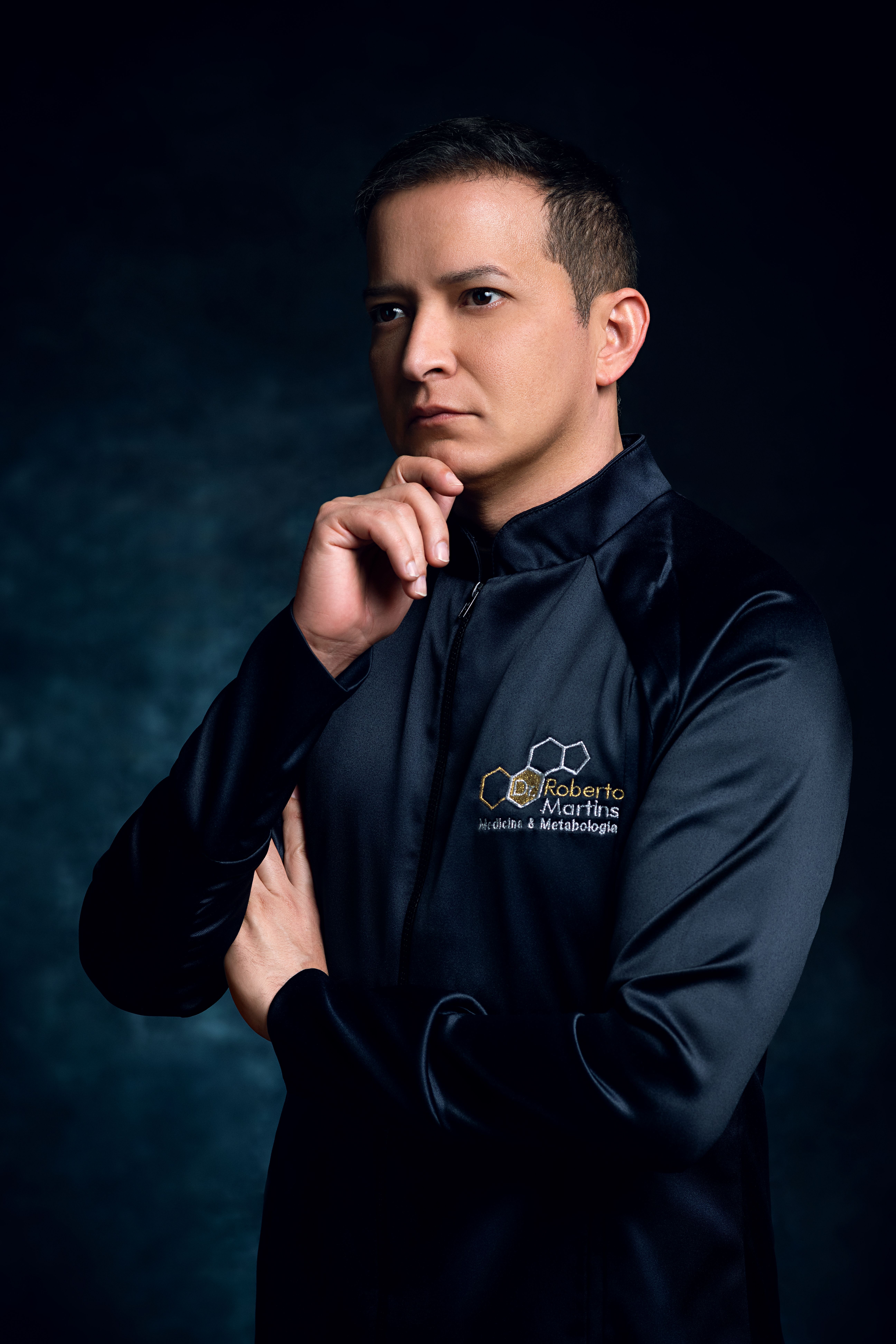 A thoughtful man in a sleek black jacket poses against a dark background.