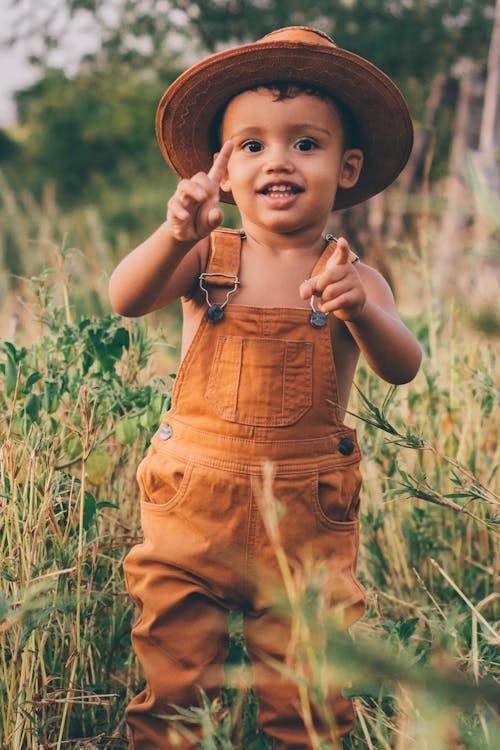 A Little Child in Overalls and Hat Standing on a Meadow in Summer 
