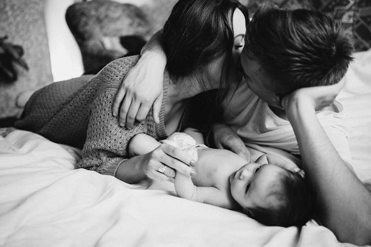 Parents Lying On The Bed With Their Newborn Baby And Kissing 