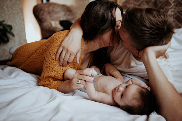 Parents Lying On The Bed With Their Newborn Baby And Kissing 