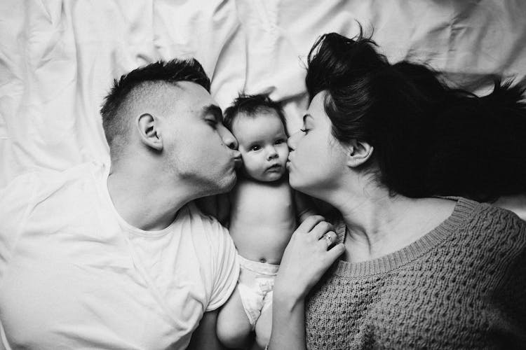 Black And White Photo Of Couple Kissing Baby Lying Between Them