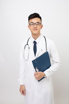 A young male doctor in a white coat stands smiling with a stethoscope and folder, isolated on white.