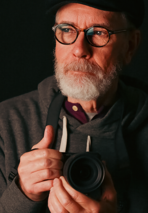 Free Elderly man wearing glasses and a cap, holding a camera, looking away thoughtfully. Stock Photo