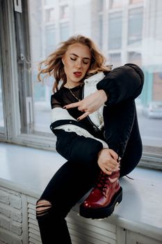 Fashionable young woman poses indoors on a city windowsill with trendy outfit and confident expression.