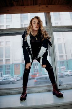 Dynamic fashion portrait of a young woman posing by a windowsill in an urban environment.