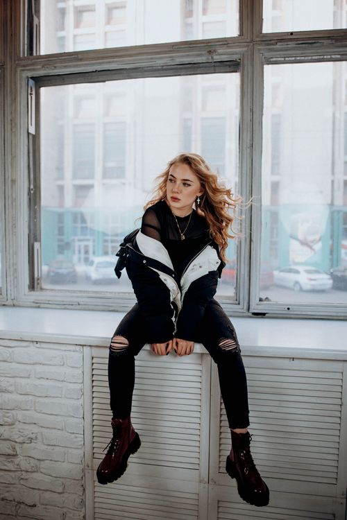 Free Young woman in trendy outfit posing on windowsill with urban backdrop, showcasing modern fashion. Stock Photo