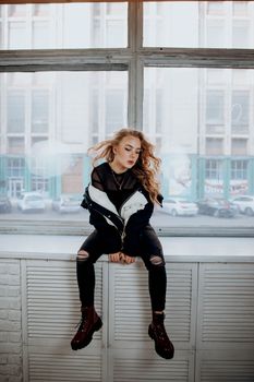 Stylish woman in trendy outfit sitting on windowsill with urban backdrop.