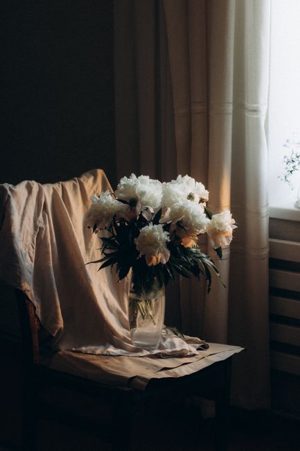 A serene arrangement of white flowers in a vase on a draped chair by a softly lit window.
