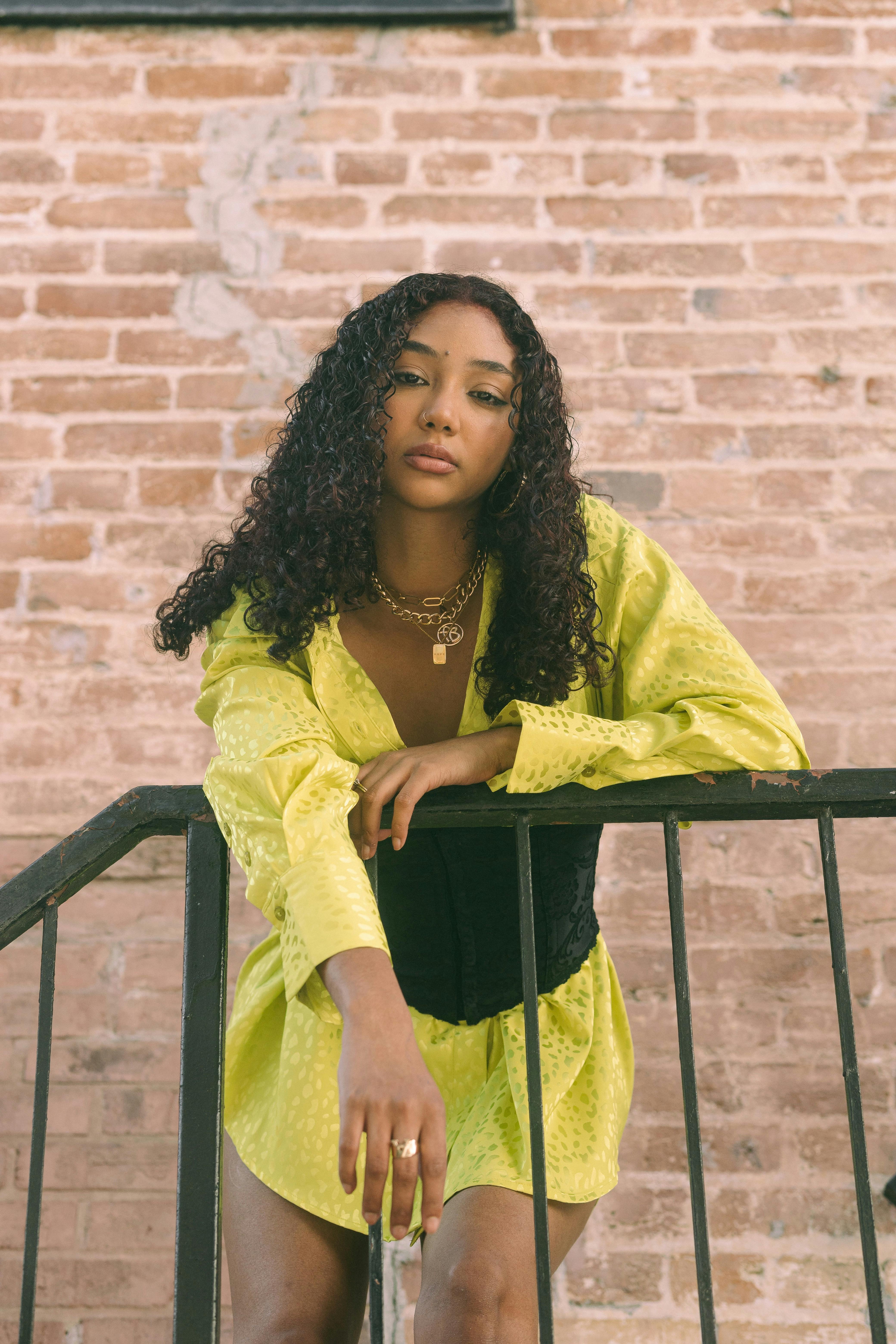 Stylish woman in a yellow dress leaning on a railing with a brick wall background.