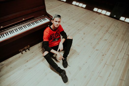 Teen sitting casually by a piano in a studio setting, expressing youthful style and attitude.