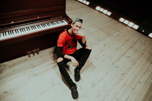 A smiling young man in casual attire sits by a piano in a well-lit music studio.