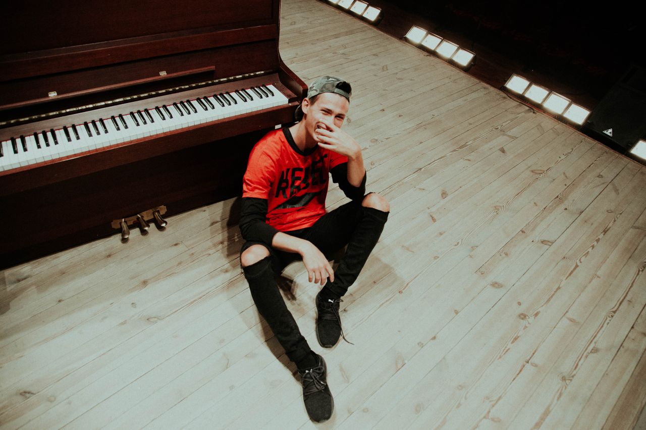 a smiling young man in casual attire sits by a piano in a well lit mus