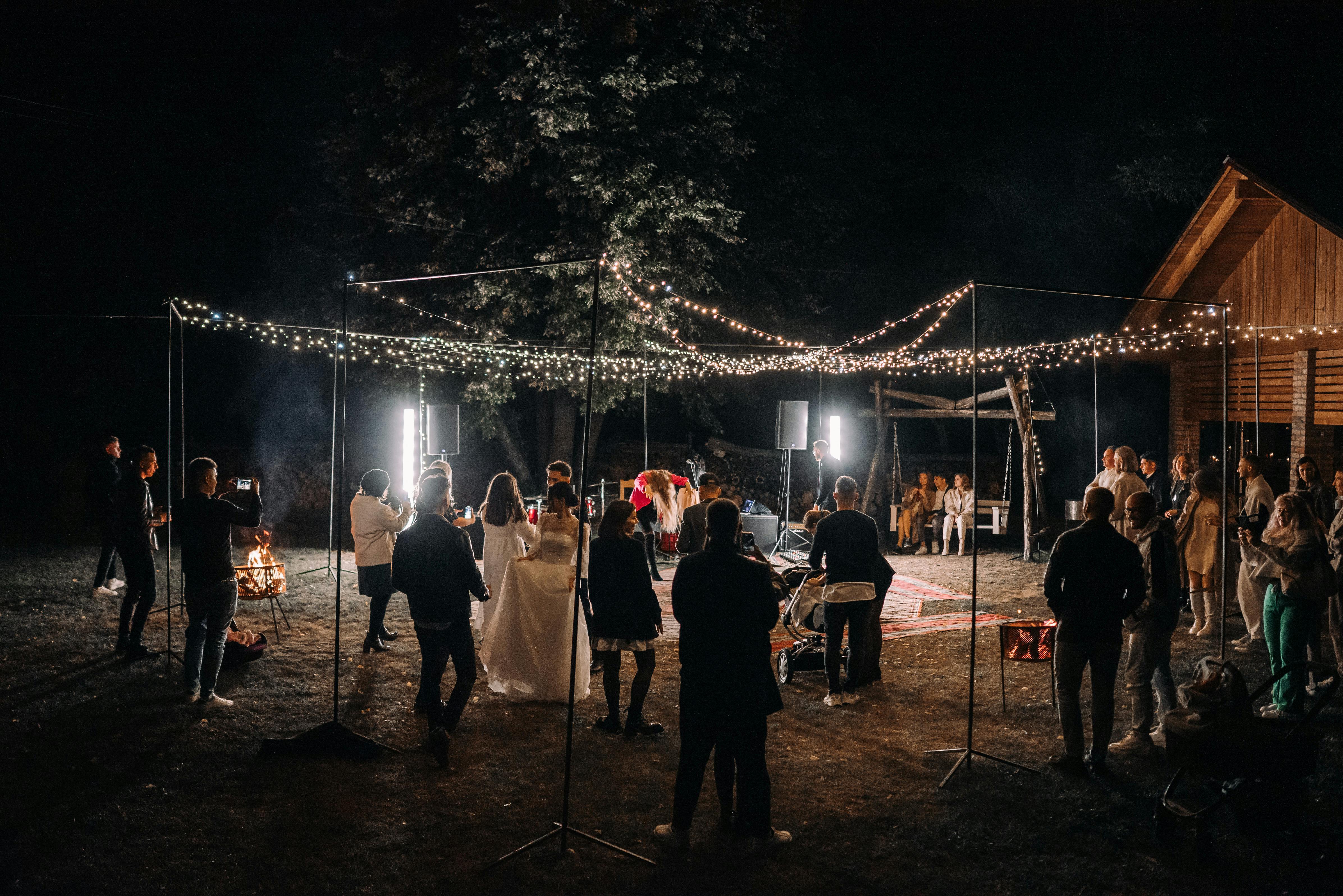 Free An outdoor wedding reception at night with string lights and guests enjoying the festivities. Stock Photo