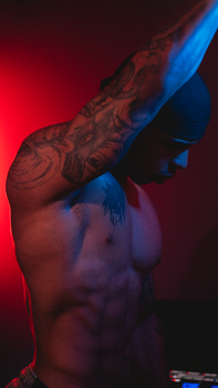 Portrait of a muscular tattooed man with arm raised, set against a vivid red background.